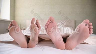一对年轻夫妇的脚从卧室的被子下伸出来。 两只赤脚互相抚摸